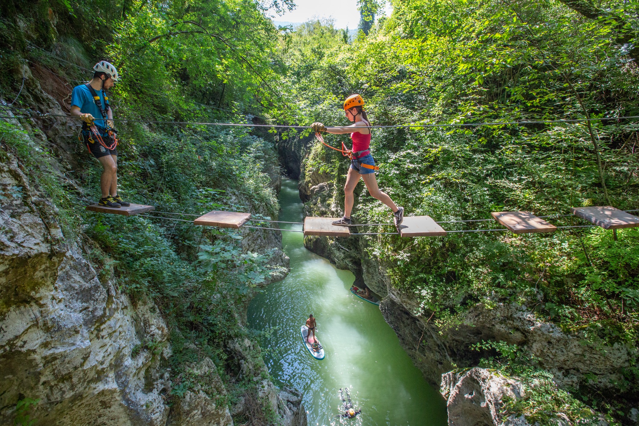 Participantes con casco y arnés Petzl cruzan un recorrido de plataformas suspendidas sobre un río en un parque de aventura de Italia.